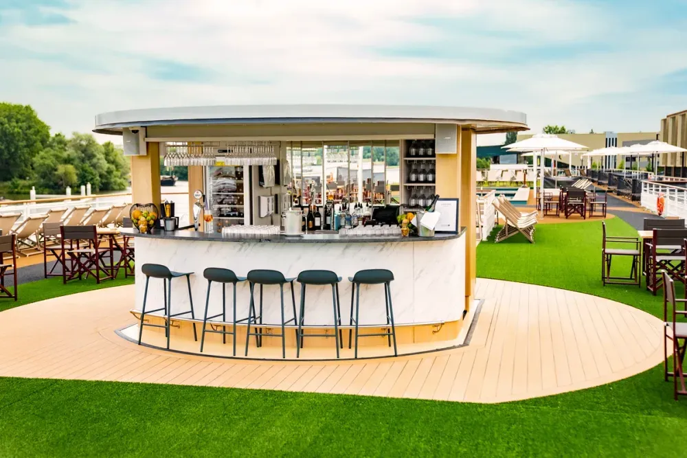 A stylish outdoor bar with a sleek marble counter is surrounded by five dark stools on a wooden deck. The bar is stocked with bottles and glasses, and fresh fruit decorates the counter. It sits on a lush green artificial lawn, with deck chairs and tables under white umbrellas in the background, creating a relaxed, sunny atmosphere.
