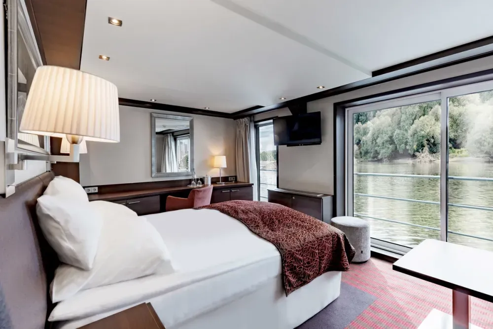 A cozy, modern bedroom features a plush bed with white pillows and a maroon patterned throw, a wall-mounted TV, and a large window offering a scenic river view.