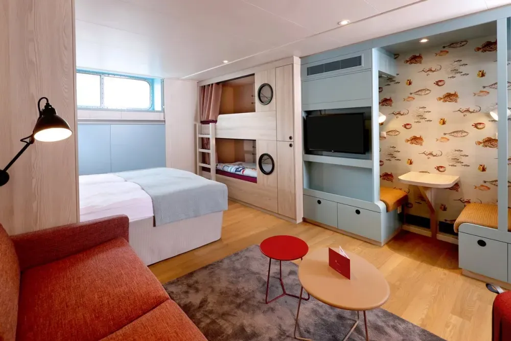 A modern, cozy cabin features a double bed with a light blue throw, a bunk bed with round portholes, and a small seating area with a TV. The wall behind the TV is decorated with whimsical sea creature wallpaper. A red sofa and two small tables, one red and one beige, add a touch of color to the room.