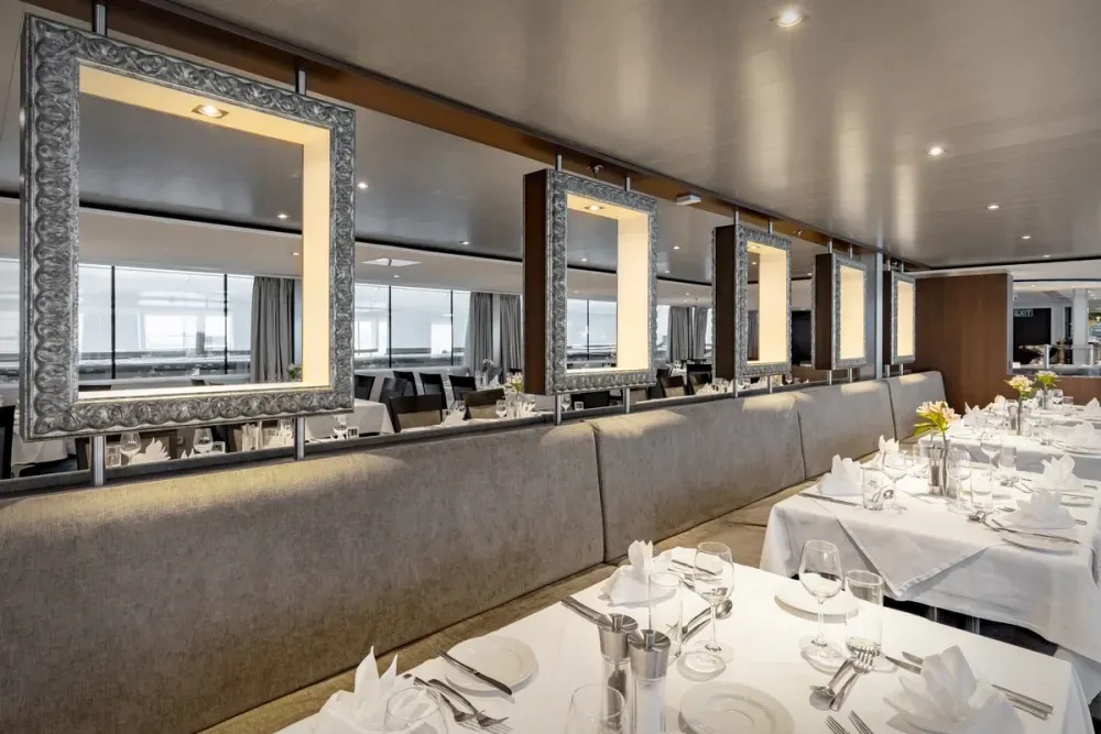 A modern, elegant restaurant interior features neatly set tables with white tablecloths, folded napkins, and glassware. Decorative frames with soft lighting hang above a long cushioned bench, creating a sophisticated ambiance. Large windows in the background provide ample natural light.