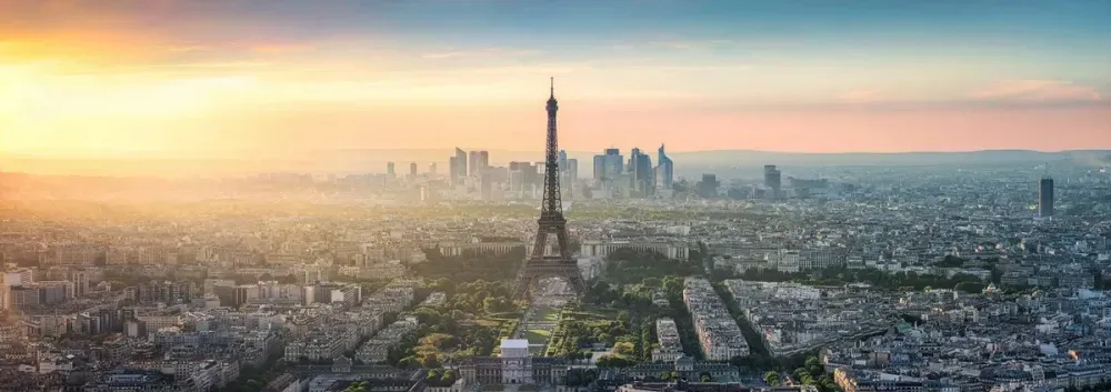 Aerial view of Paris at sunset, with the Eiffel Tower prominently in the center, surrounded by sprawling cityscape and distant modern buildings under a colorful sky.