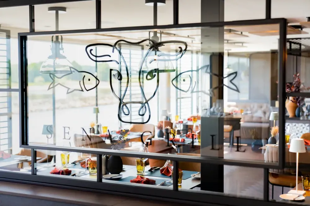 A glass window features a stylized design of a cow's head flanked by two fish, with the words "SENA GRILLE" below. Through the window, a modern restaurant interior is visible, with neatly set tables, orange napkins, and warm lighting.