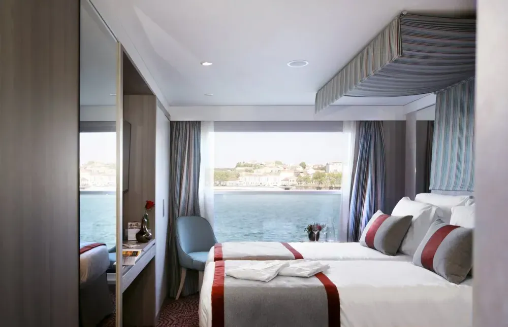 A cozy cruise ship cabin features a neatly made bed with red and gray accents, a large window offering a scenic view of the water and distant shoreline, and a small desk with a chair and decorative vase.