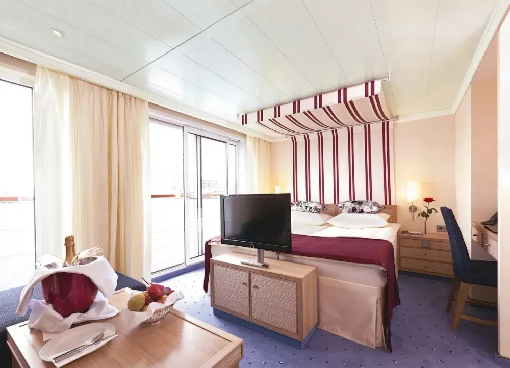 A cozy hotel room features a double bed with a burgundy and white striped canopy and matching bedding. A flat-screen TV sits on a wooden cabinet facing the bed. Large sliding glass doors with light curtains lead to a balcony. A small table holds a champagne bottle in an ice bucket and a fruit basket. A blue chair is placed at a desk in the corner, and a single red rose decorates the bedside table.