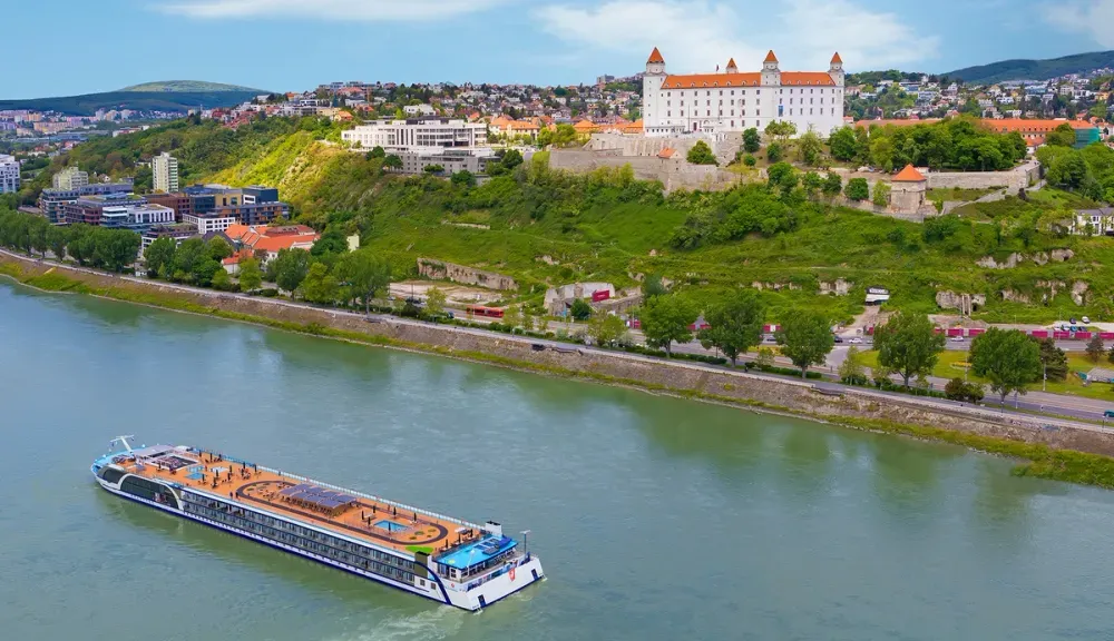 A river cruise ship sails along a wide river, with a historic castle featuring red roofs perched on a green hill in the background. The cityscape includes various buildings nestled among lush greenery, under a partly cloudy sky.