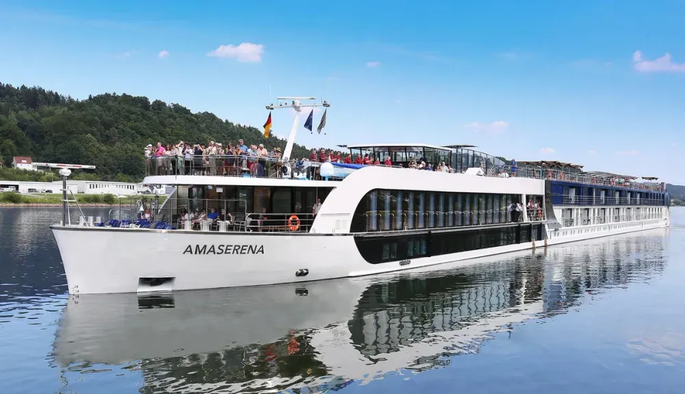 The AMA Serena cruise ship glides along a calm river, its sleek white exterior reflecting in the water. Passengers gather on the upper deck, enjoying the scenic view under a clear blue sky, with lush green trees lining the riverbank.