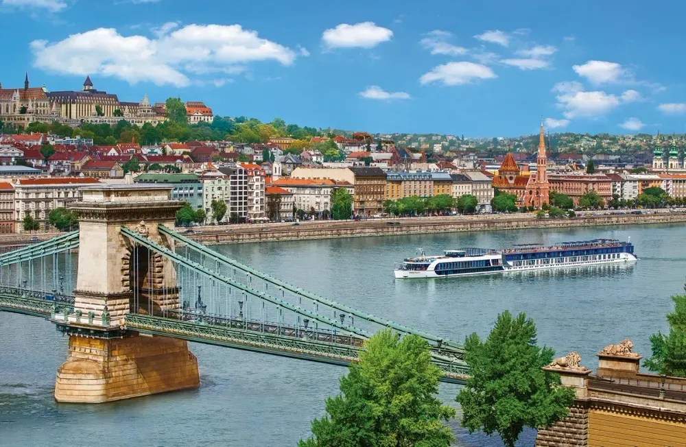 A scenic view of Budapest featuring the Chain Bridge spanning the Danube River, with a sleek river cruise boat passing underneath. The background showcases a vibrant cityscape with colorful historic buildings and a prominent church spire, all under a bright blue sky with fluffy clouds.