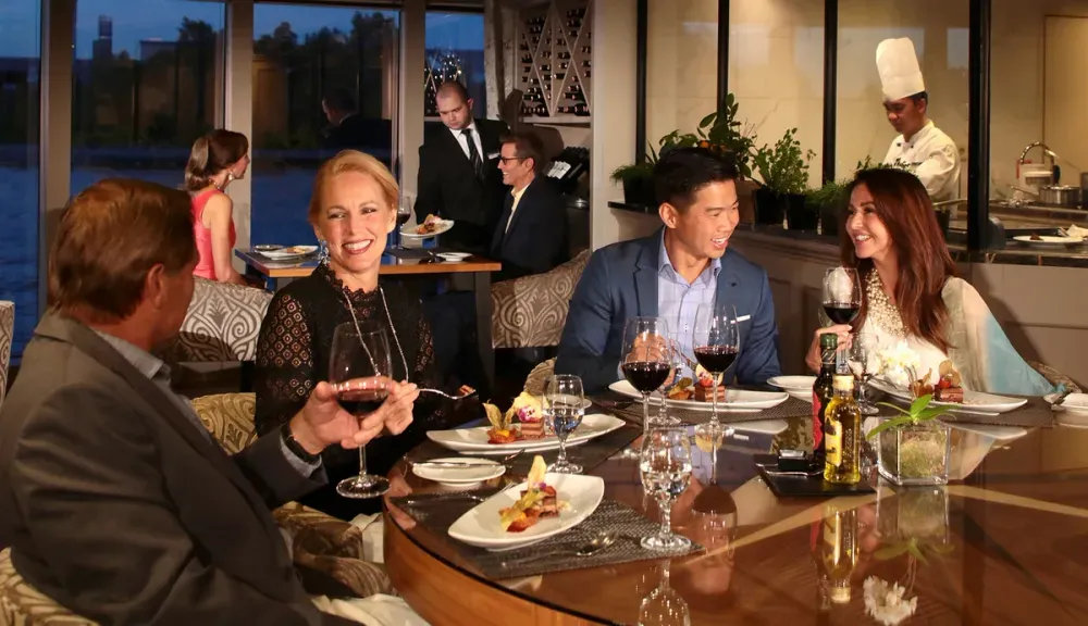 A group of four people enjoy a meal at a restaurant, seated around a table with elegantly plated dishes and wine glasses. The setting is refined, with a chef visible in the background through a glass partition. The diners are engaged in lively conversation, creating a warm and inviting atmosphere.