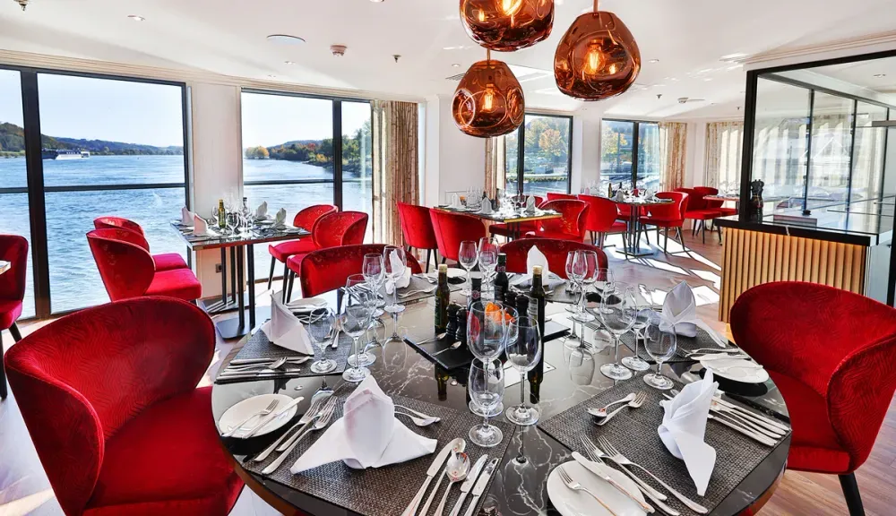 A luxurious dining area features elegantly set tables with wine glasses and folded napkins, surrounded by plush red chairs. Large windows offer a scenic view of a river, while modern pendant lights hang overhead, creating a warm ambiance.