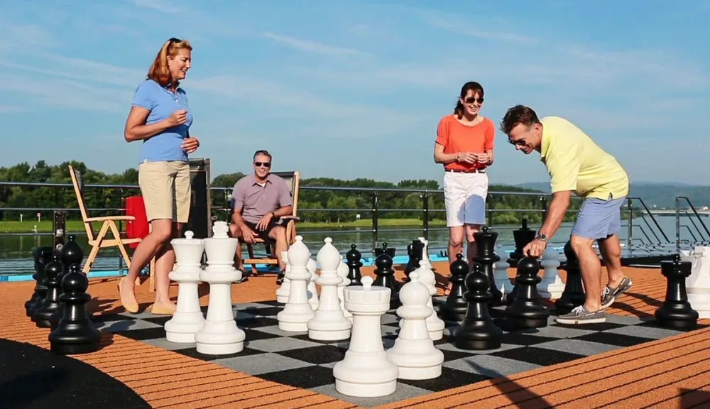 Four people are enjoying a game of giant chess on a sunny deck, with a river and lush greenery in the background. One person is making a move with a large black piece, while others watch and laugh, creating a lively and fun atmosphere.