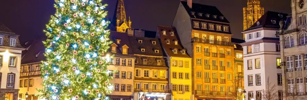 A beautifully lit Christmas tree adorned with white and blue ornaments stands prominently in a charming European square, surrounded by historic, warmly illuminated buildings with steeply pitched roofs under a night sky.