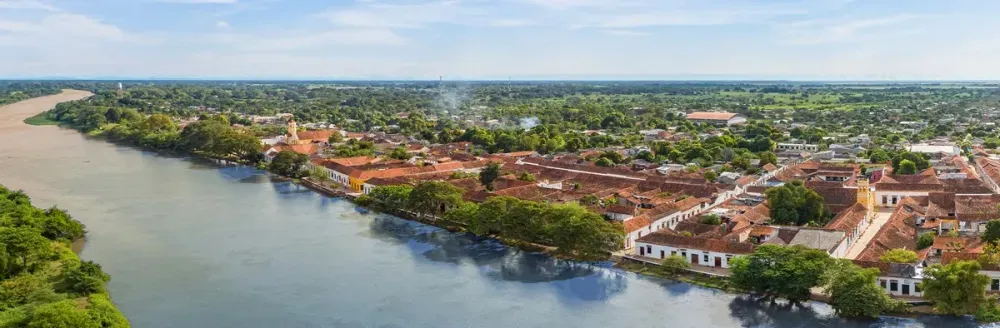 A picturesque aerial view of a quaint riverside town with red-tiled roofs nestled among lush greenery, bordered by a calm river on the left, under a clear blue sky.