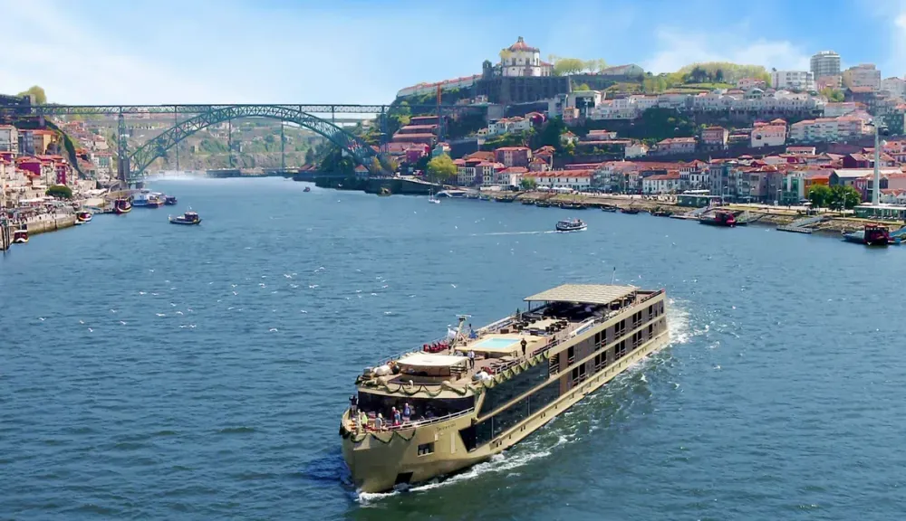 A large river cruise ship sails along a wide river, with a picturesque cityscape in the background featuring colorful buildings and a prominent arched bridge. The sun shines brightly, highlighting the vibrant scenery and clear blue sky.
