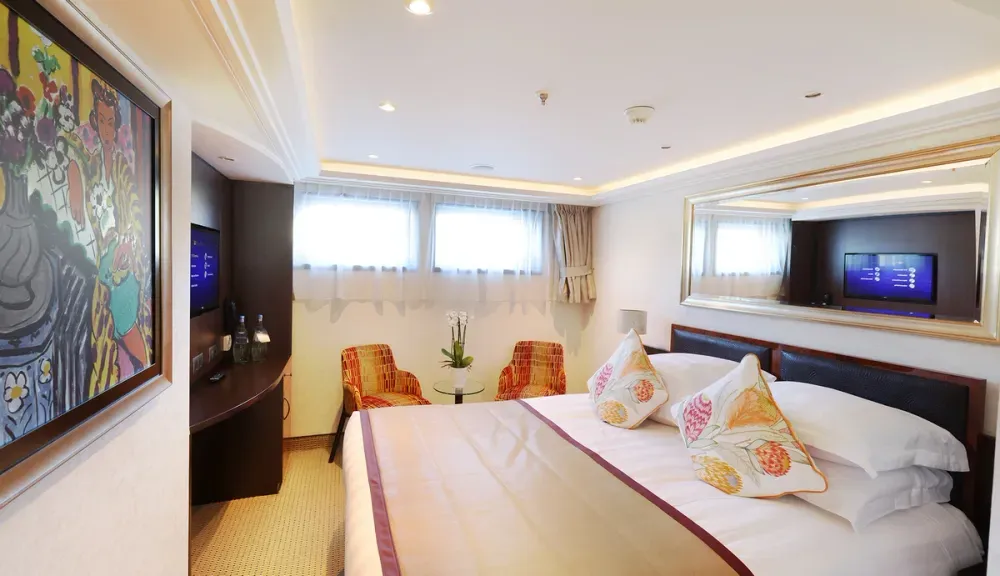 A cozy hotel room features a neatly made bed with floral-patterned pillows and a large mirror above the headboard. Two colorful chairs sit by a small table with a plant, and a TV is mounted on the wall. Soft lighting and artwork add a warm touch to the space.