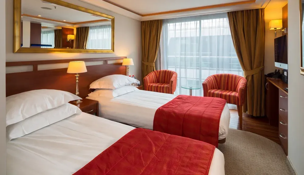 A cozy hotel room features two neatly made twin beds with white linens and red blankets. A large mirror hangs above the beds, reflecting soft light from two bedside lamps. Two striped armchairs and a small glass table sit by a large window with drawn curtains, creating a warm and inviting atmosphere.