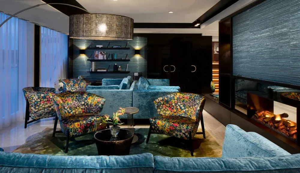 A modern living room features vibrant floral armchairs and a plush blue sectional sofa. A large, textured black pendant light hangs above, illuminating the space. The room includes a cozy electric fireplace and a wall-mounted bookshelf with decorative items, creating a stylish and inviting atmosphere.