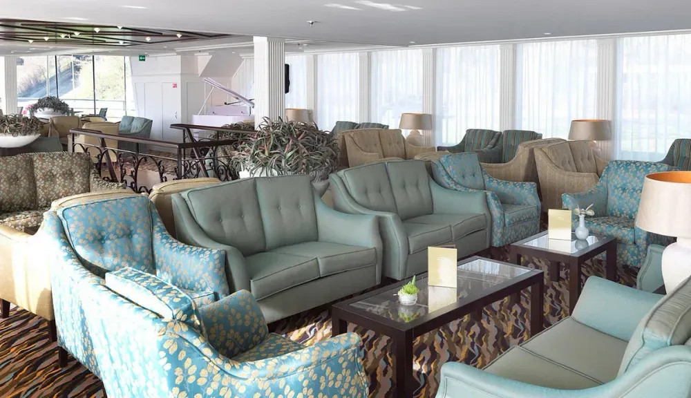 A spacious and elegant lounge area features an array of plush armchairs and sofas in soft green and patterned upholstery. The room is filled with natural light from large windows, and several small tables with decorative plants and lamps are scattered throughout. In the background, a white grand piano adds a touch of sophistication.