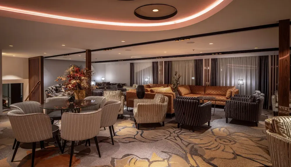 A luxurious lounge area features plush seating with striped and patterned chairs and sofas, arranged around a glass table with a decorative floral centerpiece. The space is warmly lit with soft overhead lighting, and the carpet displays an elegant floral design.