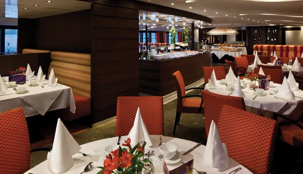 A sophisticated restaurant interior features elegantly set tables with white tablecloths and neatly folded napkins. The room is adorned with red-patterned chairs and booth seating, while a buffet area is visible in the background. Warm lighting and floral centerpieces add to the inviting atmosphere.