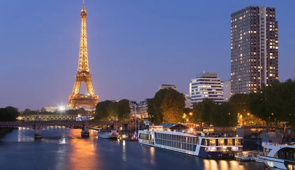 The Eiffel Tower is beautifully illuminated at dusk, reflecting on the calm waters of the Seine River, with boats docked nearby and modern buildings in the background.