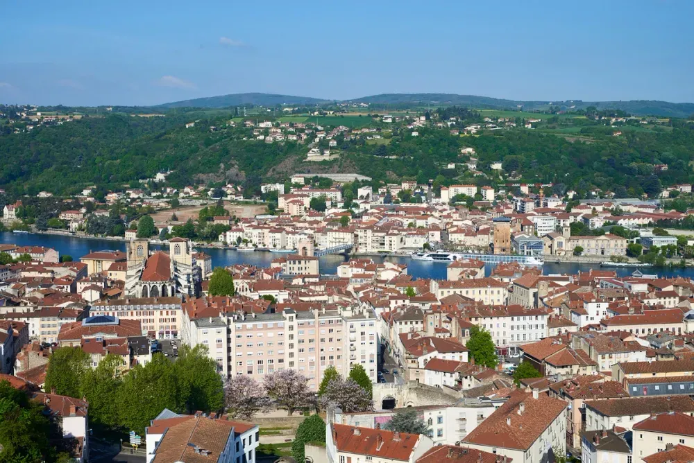 A scenic view of a city with red-tiled roofs and a prominent church near a river, surrounded by lush green hills and dotted with trees, under a clear blue sky.