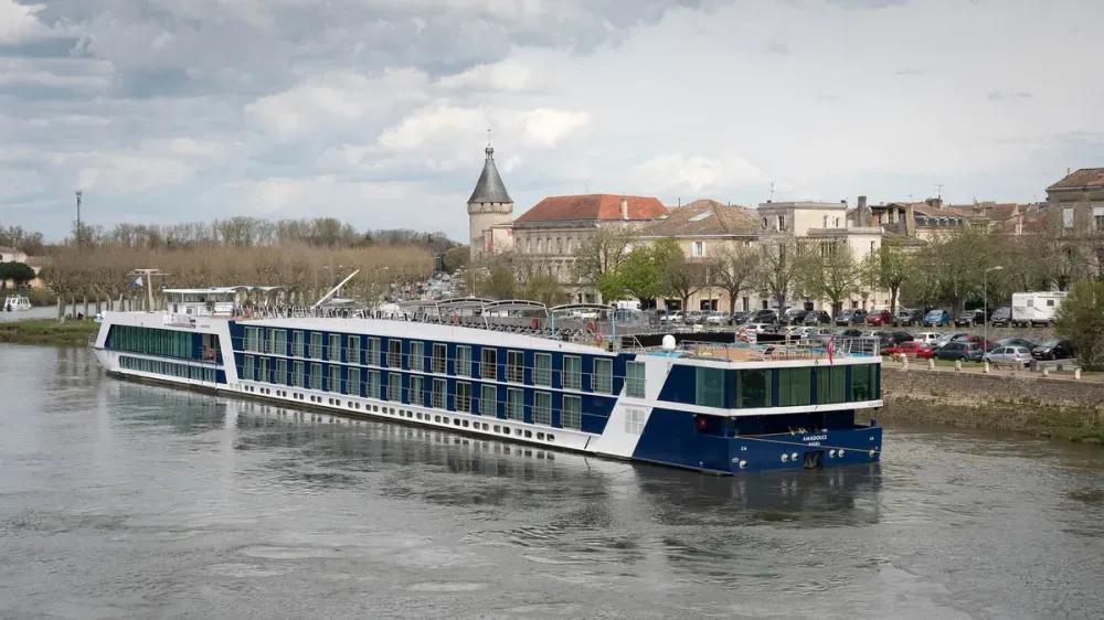 A sleek river cruise ship with a white and blue exterior is navigating a calm river, with a quaint town featuring historic buildings and a tower in the background.