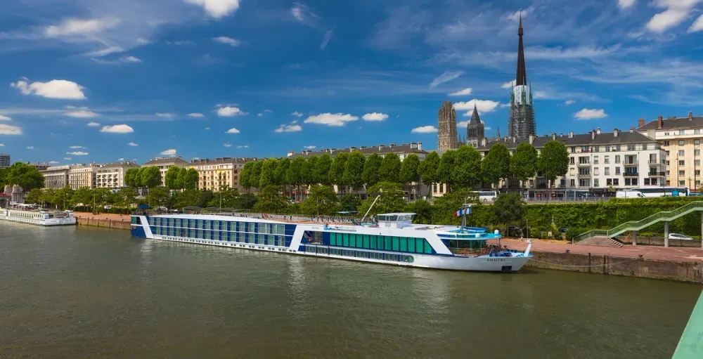 A sleek river cruise ship with a white and blue exterior is docked along a tree-lined promenade in a city. In the background, historic buildings and a tall cathedral spire rise against a vibrant blue sky dotted with fluffy clouds.