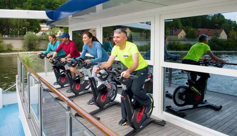 Four people are exercising on stationary bikes on a boat's deck, with a scenic river view in the background. They appear focused and are dressed in colorful athletic wear, enjoying the outdoor setting.
