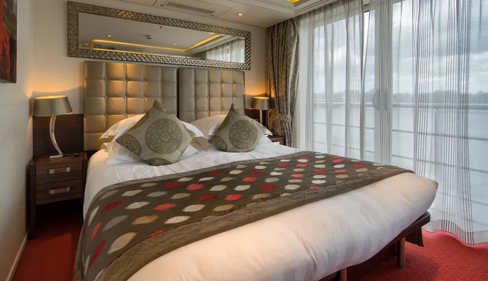 A cozy bedroom with a plush bed featuring patterned cushions and a matching throw blanket. The room has a padded headboard, a large decorative mirror above it, and is illuminated by soft lighting from bedside lamps. Floor-to-ceiling windows offer a view of a body of water, with sheer curtains partially drawn.