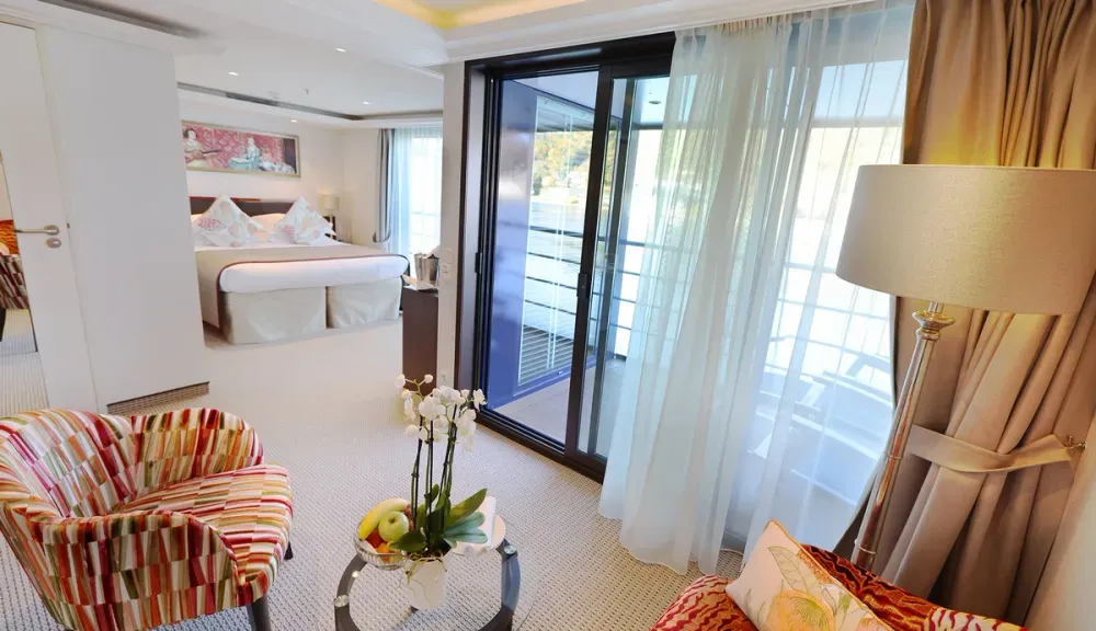 A luxurious hotel room features a cozy bed with decorative pillows and a colorful armchair. A glass sliding door leads to a balcony, allowing natural light to brighten the space. A stylish floor lamp and a table with an orchid and fruit bowl enhance the elegant ambiance.