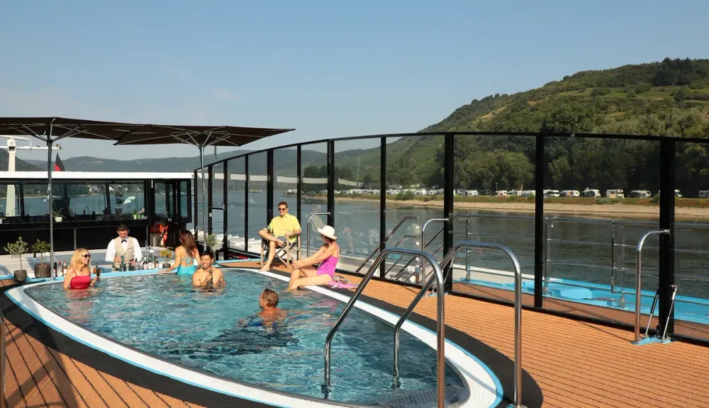 A group of people relaxes by a small pool on a cruise ship deck, enjoying drinks under a clear blue sky. The deck features modern glass railings, offering a scenic view of lush green hills and a calm river in the background.