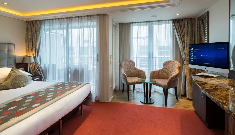A cozy hotel room features a neatly made bed with patterned pillows and a decorative runner. Two patterned chairs and a small glass table sit by large windows with sheer curtains, allowing natural light to fill the space. A flat-screen TV is mounted on a wall with a sleek, marble-topped console beneath it.