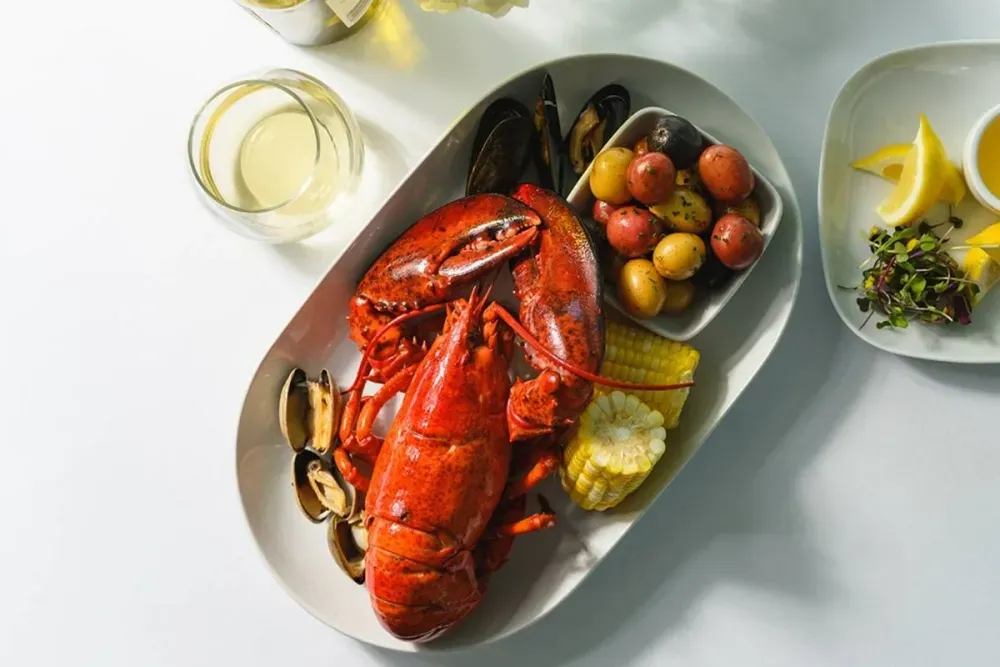 A vibrant seafood platter featuring a whole cooked lobster, clams, and mussels, accompanied by small roasted potatoes and a corn cob, is elegantly presented on a white plate. A glass of white wine sits nearby, and a separate dish holds lemon wedges and microgreens.