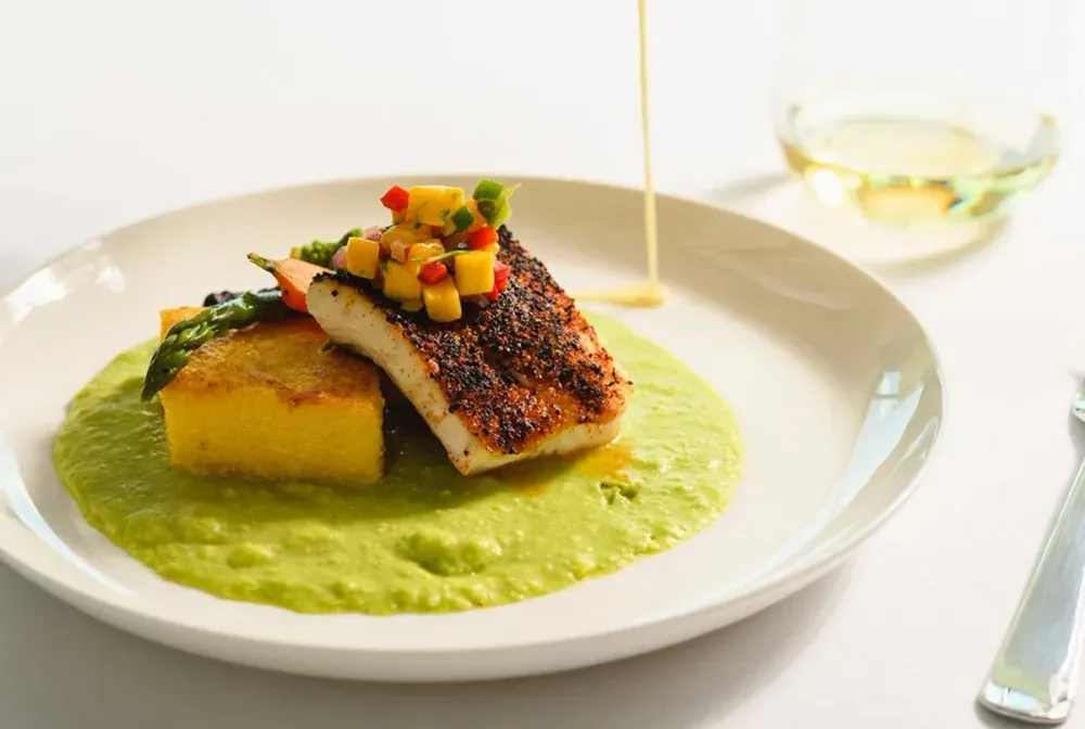 A beautifully plated dish featuring a seared piece of fish topped with a colorful mango salsa, resting on a golden polenta square and surrounded by a vibrant green sauce, with a drizzle being poured over it; a glass of white wine sits in the background.