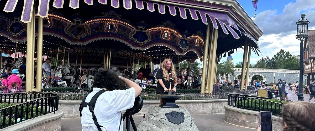 A person poses with the Sword in the Stone at a theme park, while another person takes a photo. There's a colorful carousel in the background, and several visitors can be seen enjoying the park atmosphere.