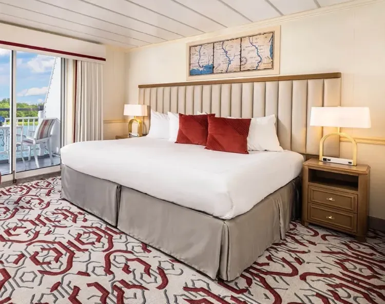 A spacious hotel room features a large bed with crisp white linens and three red accent pillows, flanked by two elegant bedside lamps on wooden nightstands. A patterned carpet in red and gray covers the floor, and a map artwork hangs above the headboard. Sliding glass doors open to a balcony with a view of greenery.