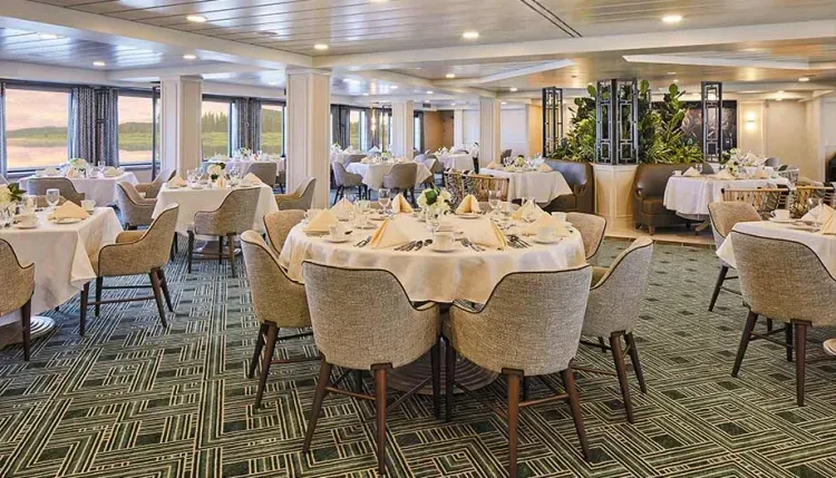 An elegant dining room features round tables set with white tablecloths, neatly arranged napkins, and tableware. Comfortable gray upholstered chairs surround the tables, and large windows offer scenic views. The room is decorated with plants and has a patterned green carpet, creating a sophisticated atmosphere.