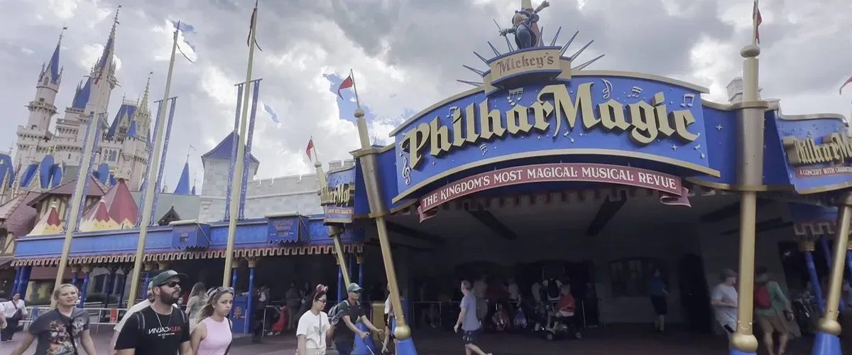 Visitors walk past the entrance to Mickey's PhilharMagic, a vibrant attraction with a blue and gold sign at a theme park, under a cloudy sky.