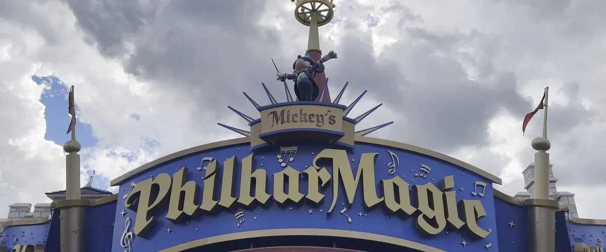 The entrance sign for "Mickey's PhilharMagic" features bold, stylized lettering with musical notes and a small character conducting from the top, set against a cloudy sky backdrop.