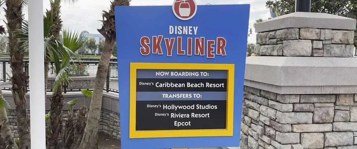 A colorful sign for the Disney Skyliner is displayed near stone and palm trees, indicating boarding for Disney's Caribbean Beach Resort and transfers to Hollywood Studios, Riviera Resort, and Epcot.