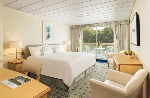 A bright, inviting hotel room features a large bed with white linens and decorative pillows. A wooden desk with a magazine sits to the left, while a comfortable armchair and a small table with a bowl of fruit are on the right. Sliding glass doors open to a balcony with chairs, offering a view of lush greenery outside.