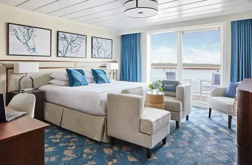 A bright and spacious hotel room features a large bed with white linens and blue accent pillows, flanked by two armchairs. The room has a nautical theme with three framed artworks above the bed and blue curtains framing a sliding glass door that opens to a balcony with a water view. A desk with a laptop and a small table with a plant add to the cozy atmosphere.