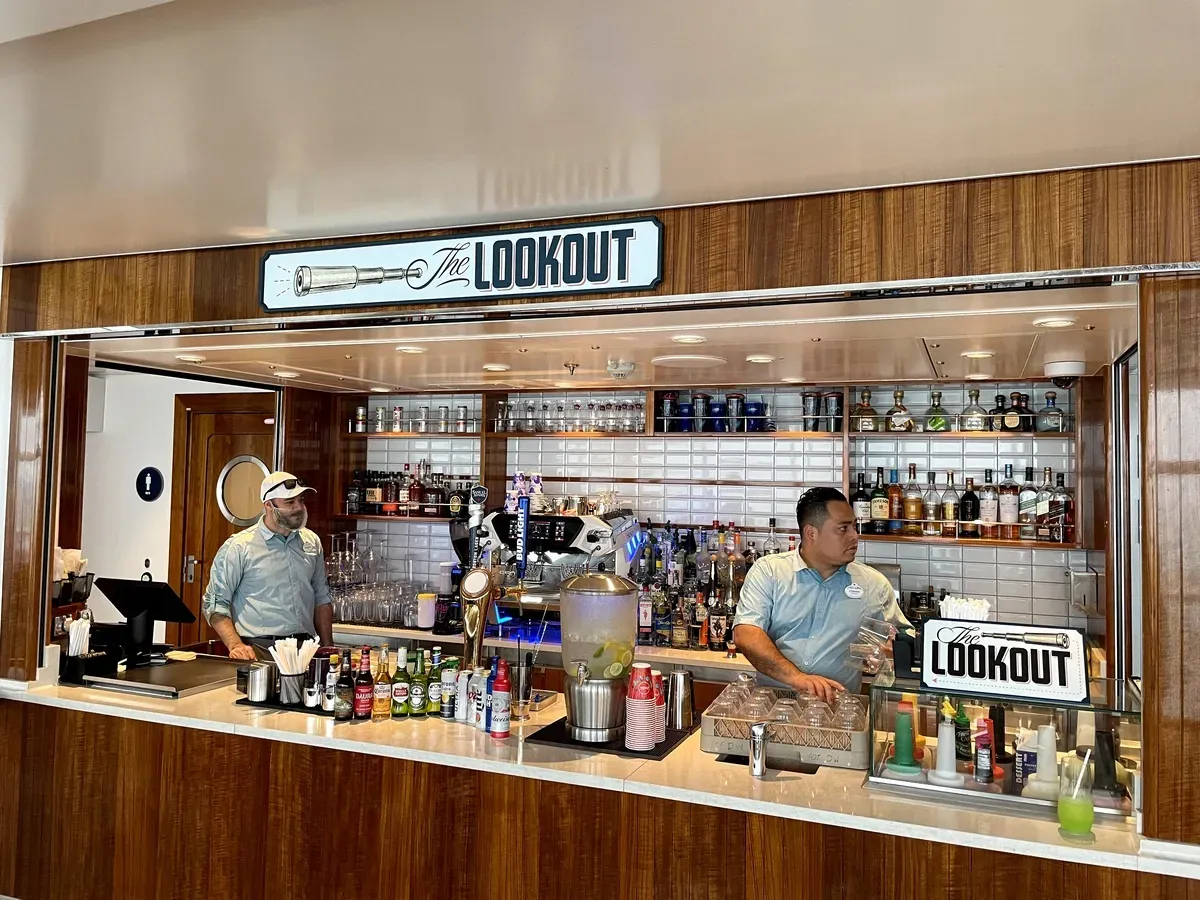 Two bartenders stand behind the polished wooden counter of "The Lookout" bar, surrounded by an array of colorful bottles and a coffee machine, with a sign displaying the bar's name above them.