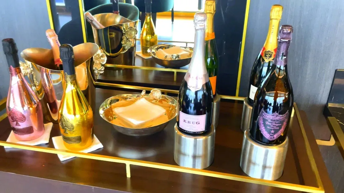 A luxurious display features several bottles of champagne, including gold and pink metallic bottles, elegantly arranged on a wooden tray with a mirrored backdrop. A decorative ice bucket and a gold dish add to the opulent setting.
