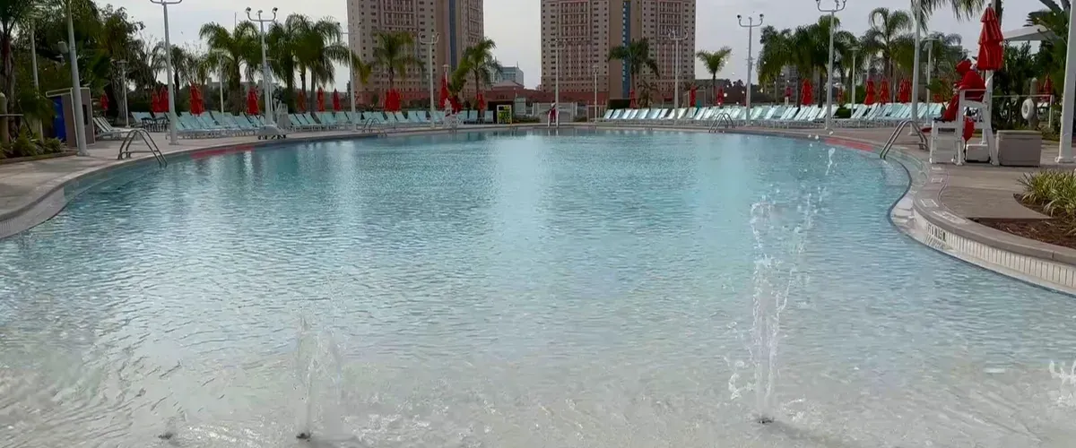 A large swimming pool with clear blue water features several small fountains near the shallow end. Surrounding the pool, numerous lounge chairs with red umbrellas provide a relaxing atmosphere, while palm trees and tall buildings are visible in the background.