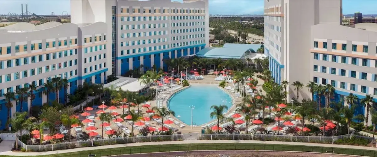 A large hotel complex featuring two tall, white buildings with blue accents surrounds a spacious, inviting swimming pool. The pool area is dotted with numerous red umbrellas and palm trees, creating a tropical atmosphere.